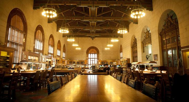 Image is of the Architecture and Planning Library Reading Room. It is a long room filled with dark furniture and lined with bookshelves and ornate doors and windows.