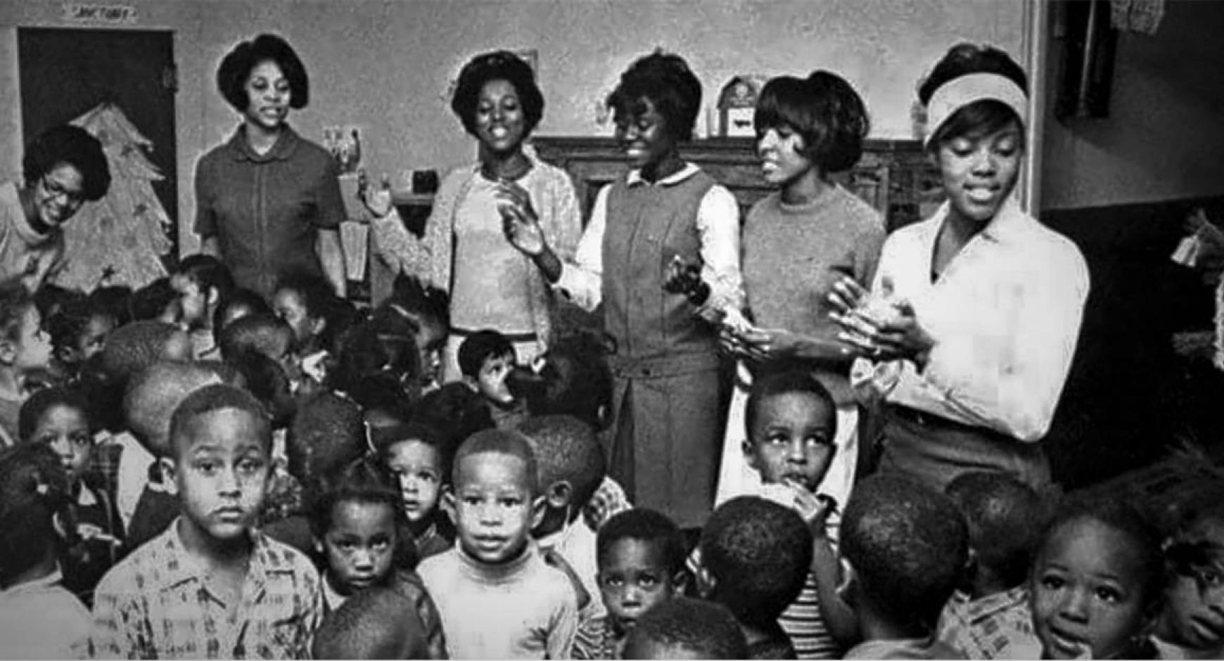The image is a black and white photo of five Alpha Kappa Alpha Sorority members, along with a large group of school children.  The women seem to be leading the children in song.  A paper Christmas tree in the background suggests this photo was taken in December, while the style of clothing worn suggests the photo is from the sixties.