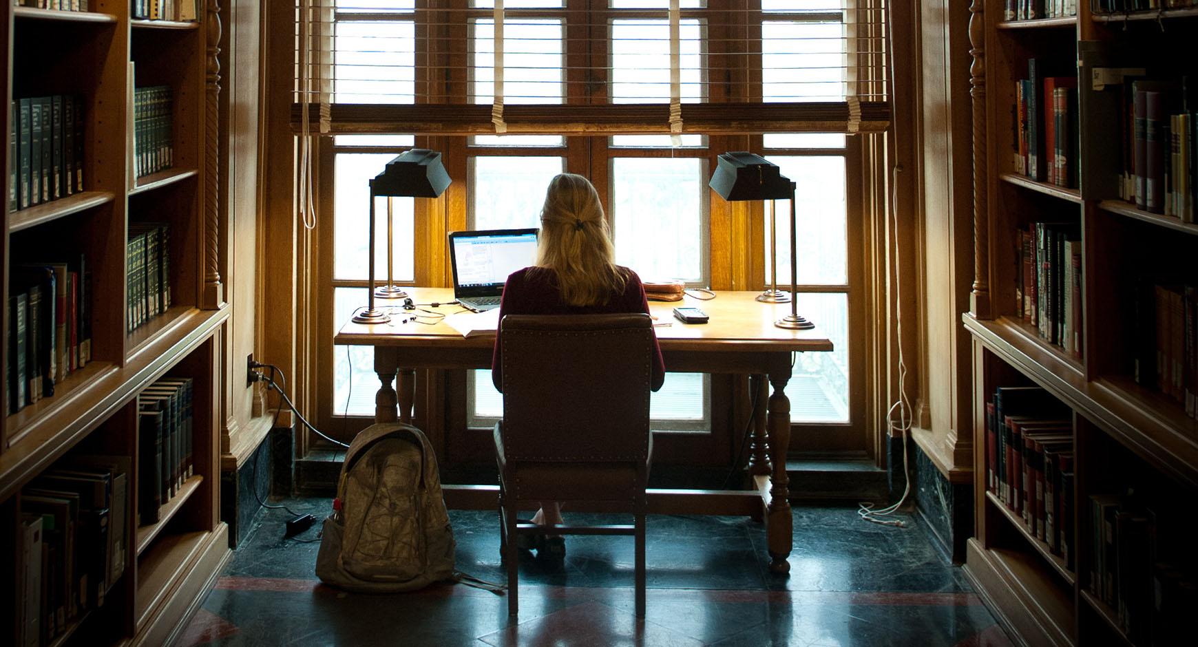 woman sitting at desk in front of window with back to camera, light streaming through, surrounded by books, working on a laptop