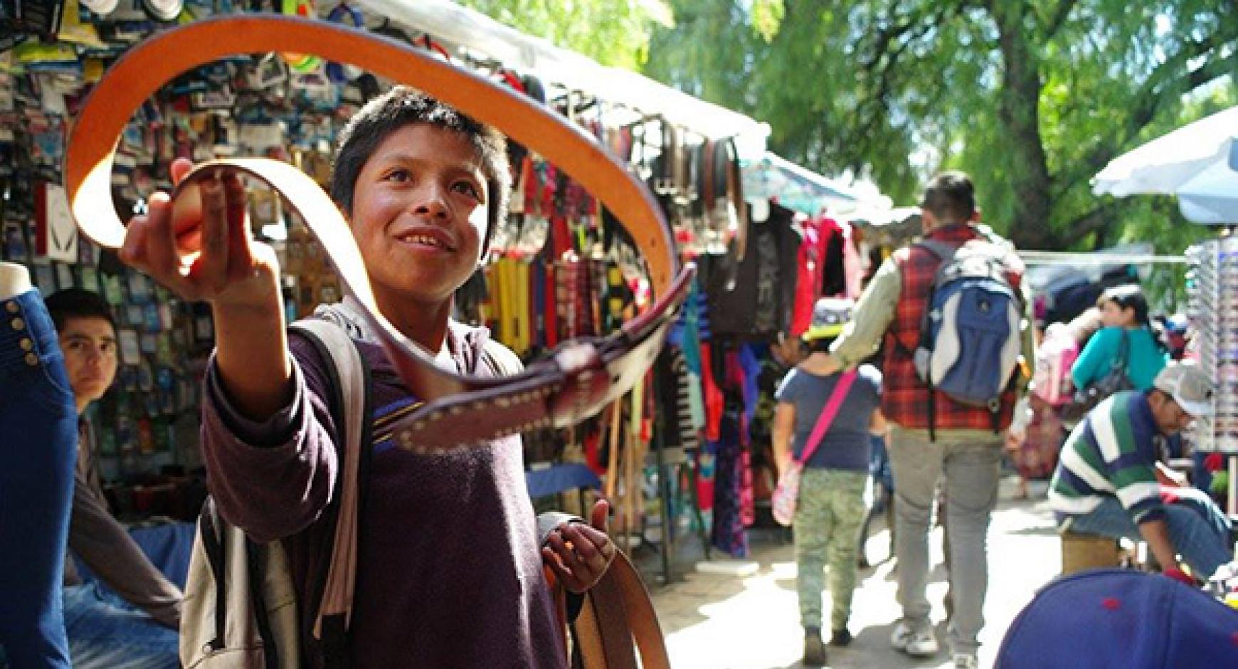 Winning photograph entitled "Cinturoncito" by Scott Squires. Image is of a smiling, young Latin-American boy standing in an outdoor market holding up a small leather belt.