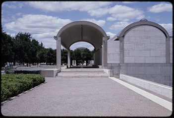 Image is an outdoor shot of the southwest exterior of the Kimbell Art Museum, Kahn Building.  It shows a stone or cement walkway, leading into to a tall, arched breezeway.  There are trees and bushes along the left side of the image. 