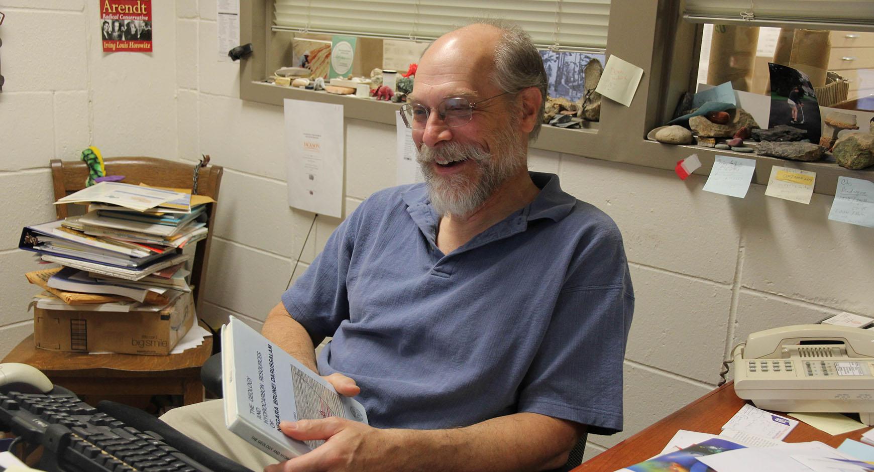 bearded man with glasses (dennis trombatore) smiling at desk with book in hands