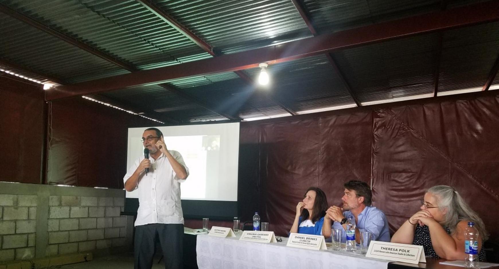 The image is of the participants/presenters sitting at a table at the AHPN seminar. The two visible name plates say Daniel Brinks and Theresa Polk. Behind the table is a screen with a slide projected onto it.  In front of the table a man stands holding a microphone and speaking.