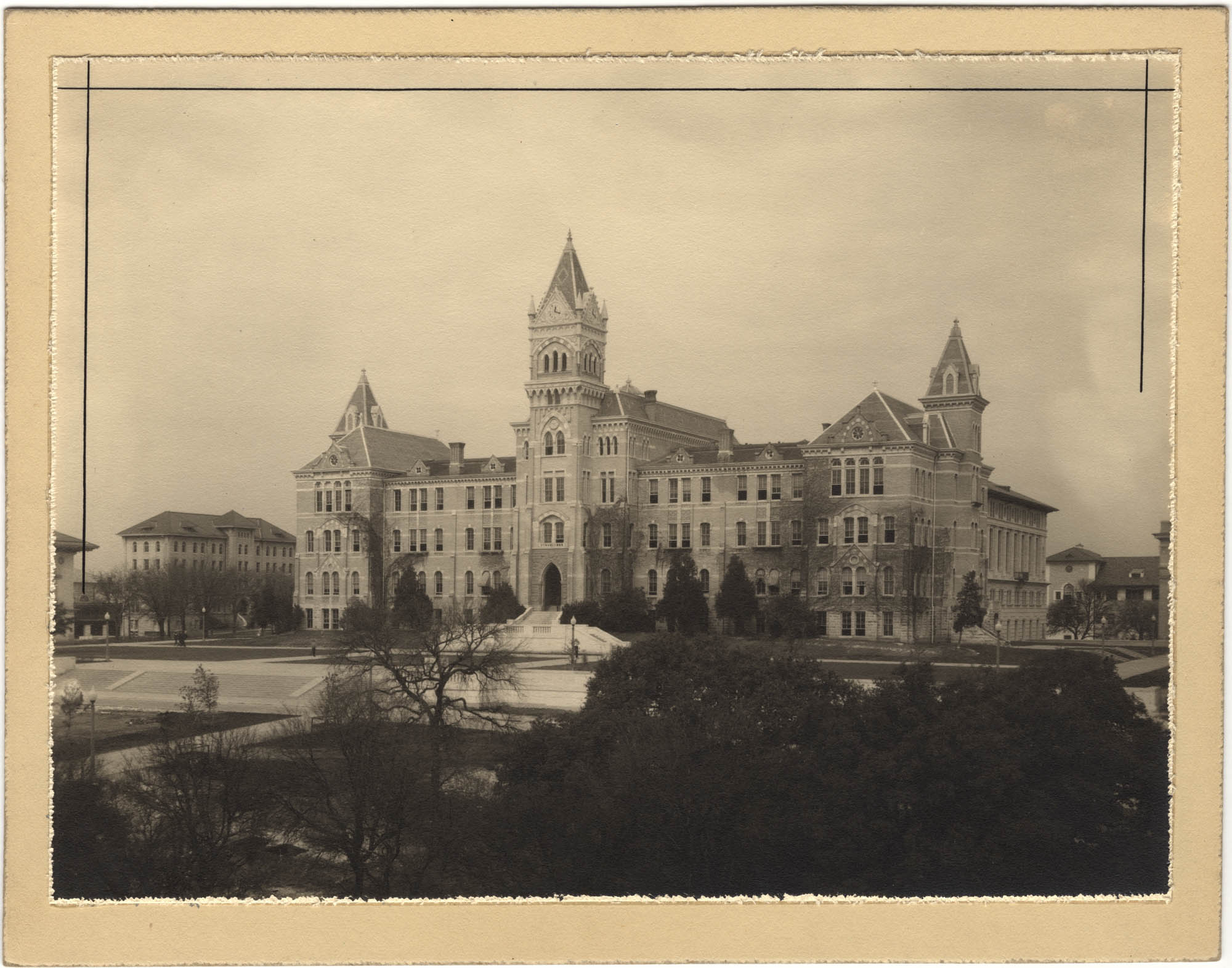 The image is a sepia photograph of the exterior of the old main building at the University of Texas.  The large building has many windows across the front that suggest four main floors.  Coming from the bottom center of the building is an elegant set of steps leading to the main door.  A tall clock tower is set at the top of the building, above the main entrance. From the Frederick Ernst Ruffini collection.