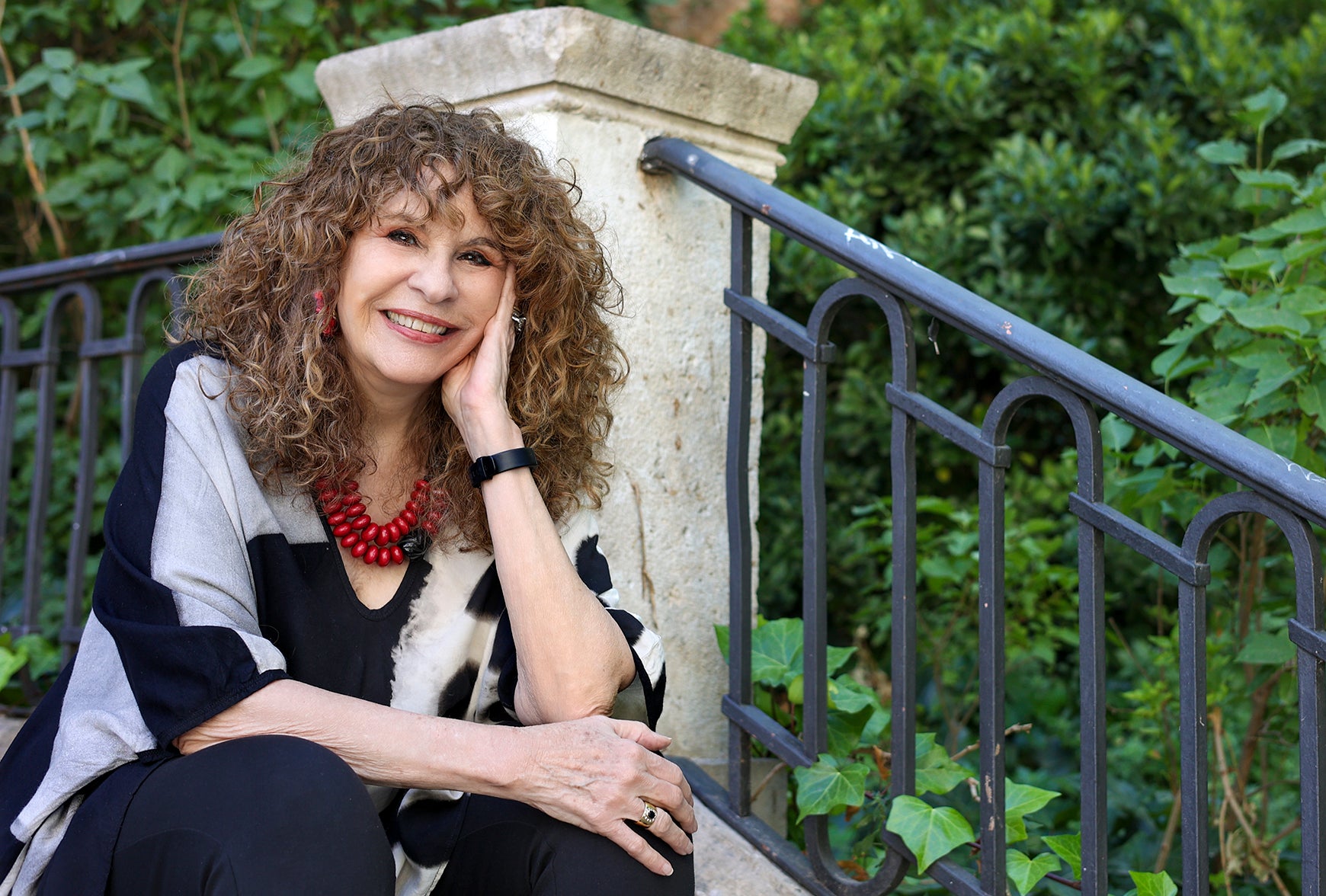 woman sitting on stone stair with iron railing leaning on hand, smiling, green plants in background.  photo by Ian Wagemann