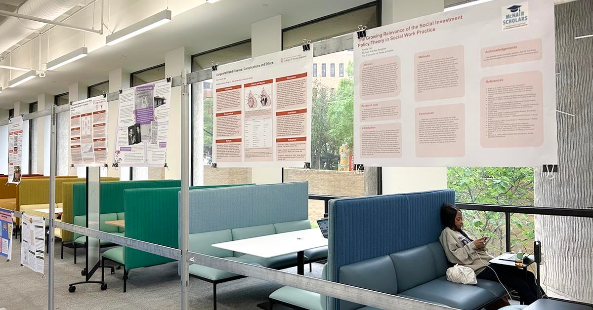 Research posters hanging on free-standing longitudinal frame in study area of scholars lab. Cafe benches in bright colors with solitary student studying and windows with a view of outdoors in the background.