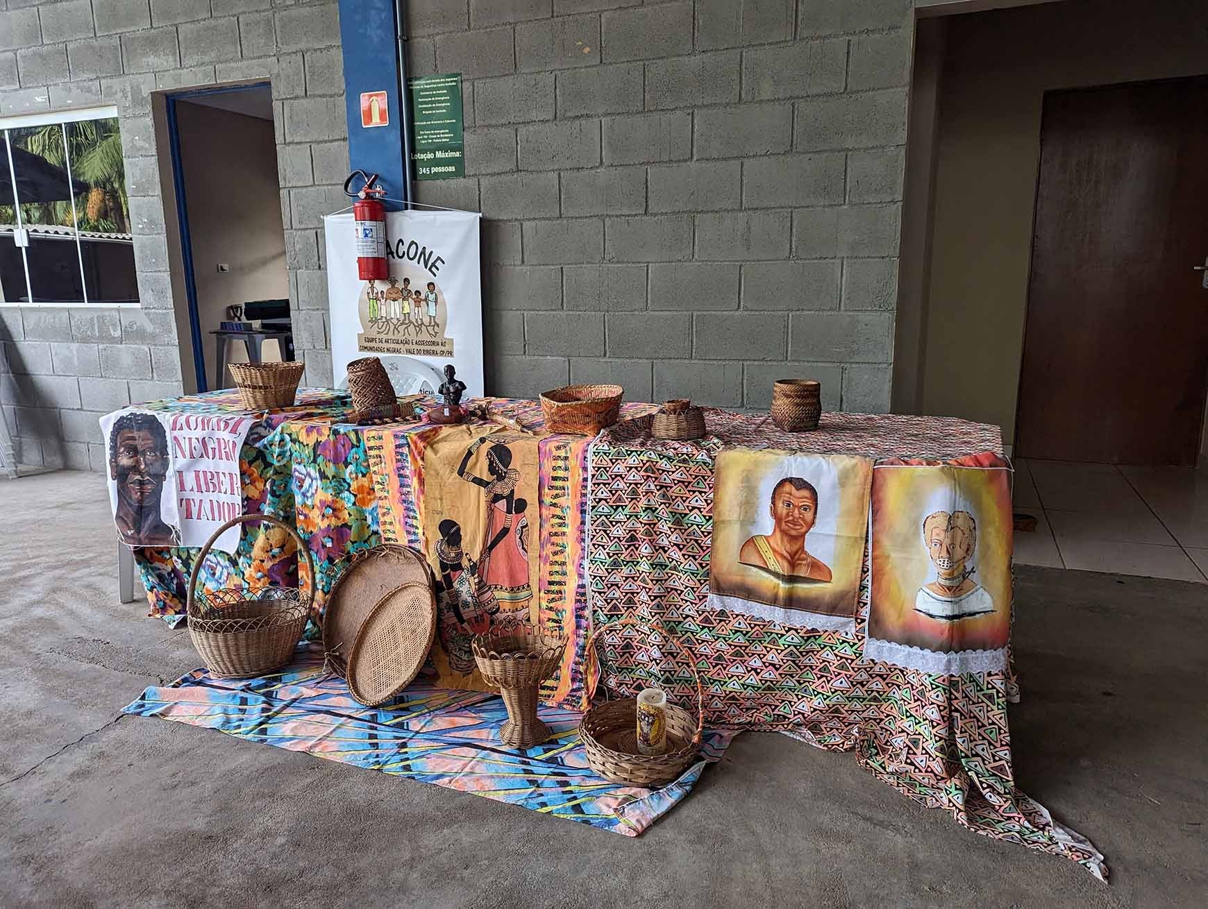 Image of a display table at a quilombo communities meetup in Eldorado, Vale do Ribeira, São Paulo, Brazil. Table draped with textile, featuring basketry and folk images.