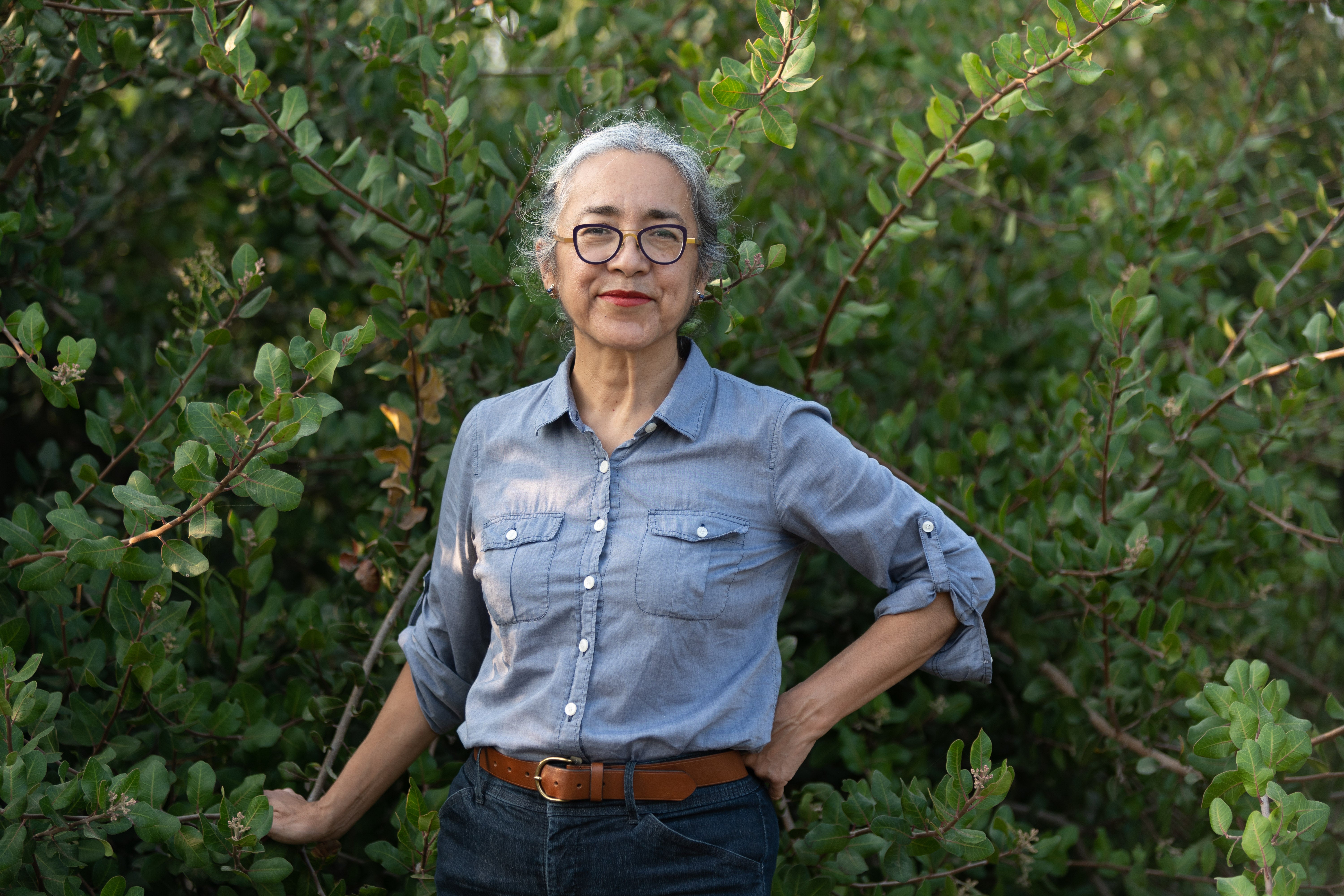 Cristina Rivera Garza wearing glasses and a blue shirt stands with hands on hips in front of green foliage.