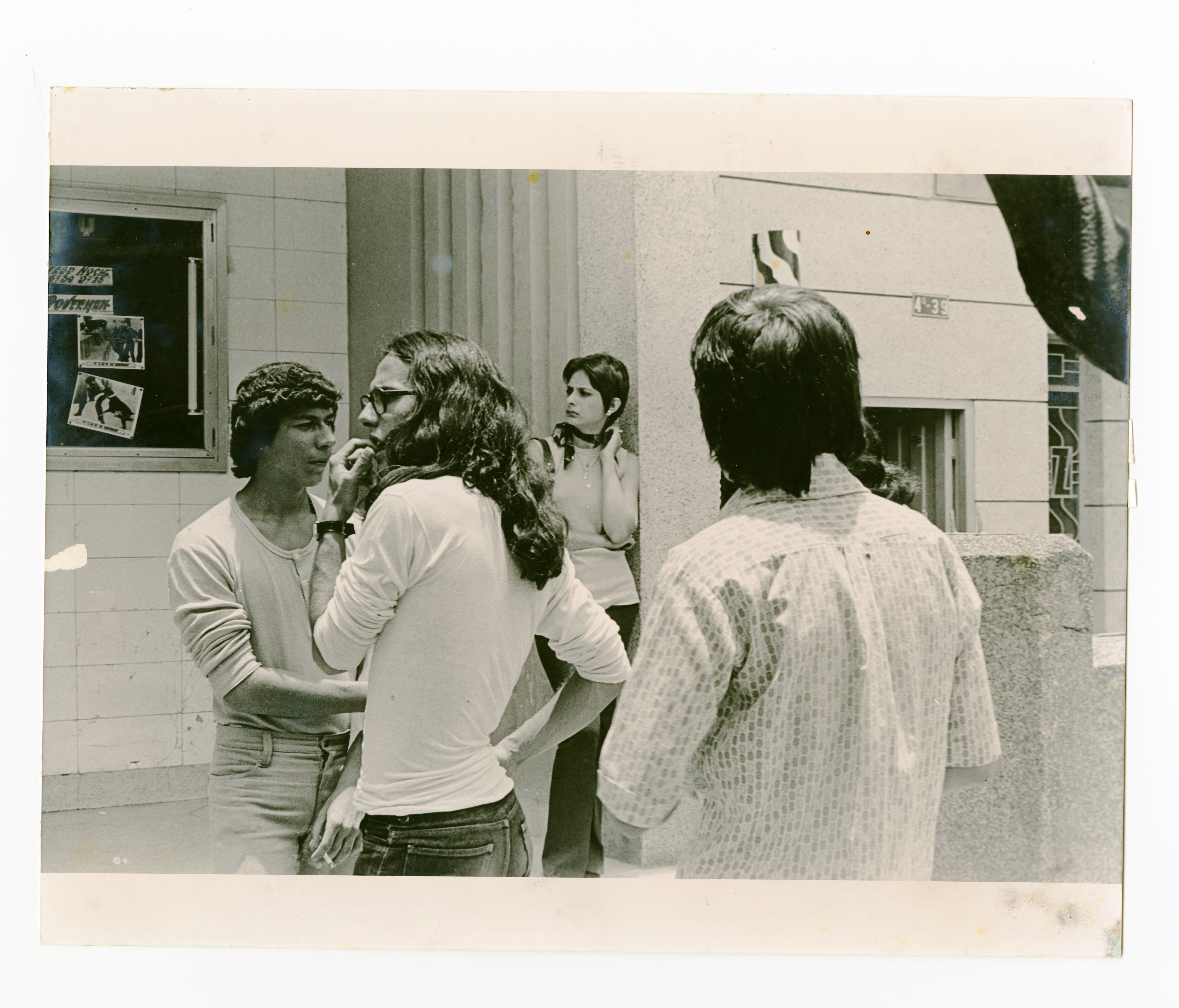A vintage black-and-white photo showing a group of four young adults gathered outside a building. In the foreground, two men face each other in conversation—one with long hair, glasses, and a hand on his hip, and the other wearing a knit cap and casual shirt. A third man, seen from behind, stands nearby, partially turned toward them. In the background, a woman leans against a wall with her hand on her neck, watching the group. Behind her, a window displays posters, including an image of a boxing match. The 