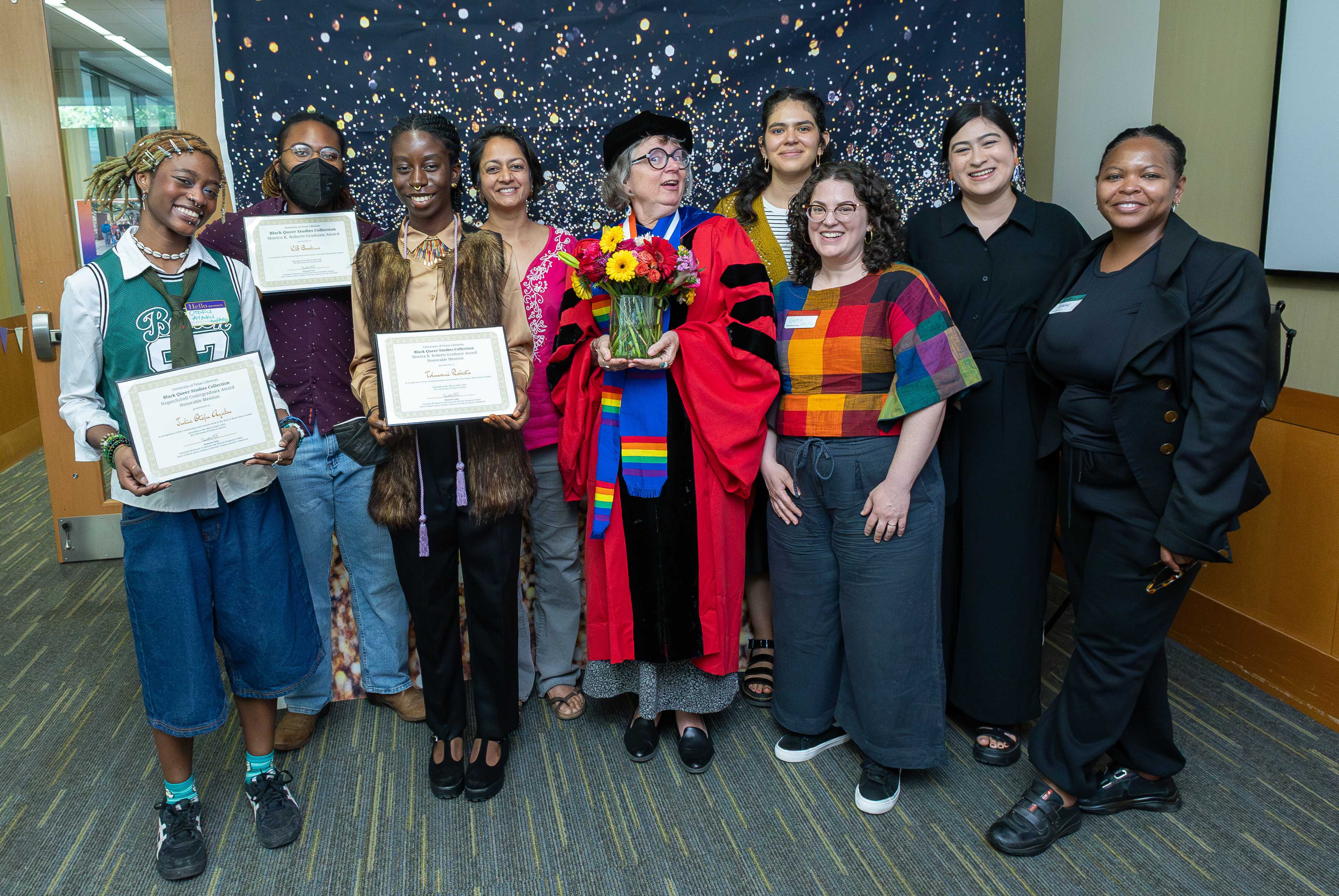 A group of ten people stands smiling in a celebratory setting. At the center, a person in academic regalia holding a bouquet of flowers is surrounded by colleagues and students. Two individuals on the left are holding framed certificates. The backdrop features a starry pattern, adding to the festive atmosphere. The group appears diverse in age, style, and background, suggesting a university or academic award ceremony.