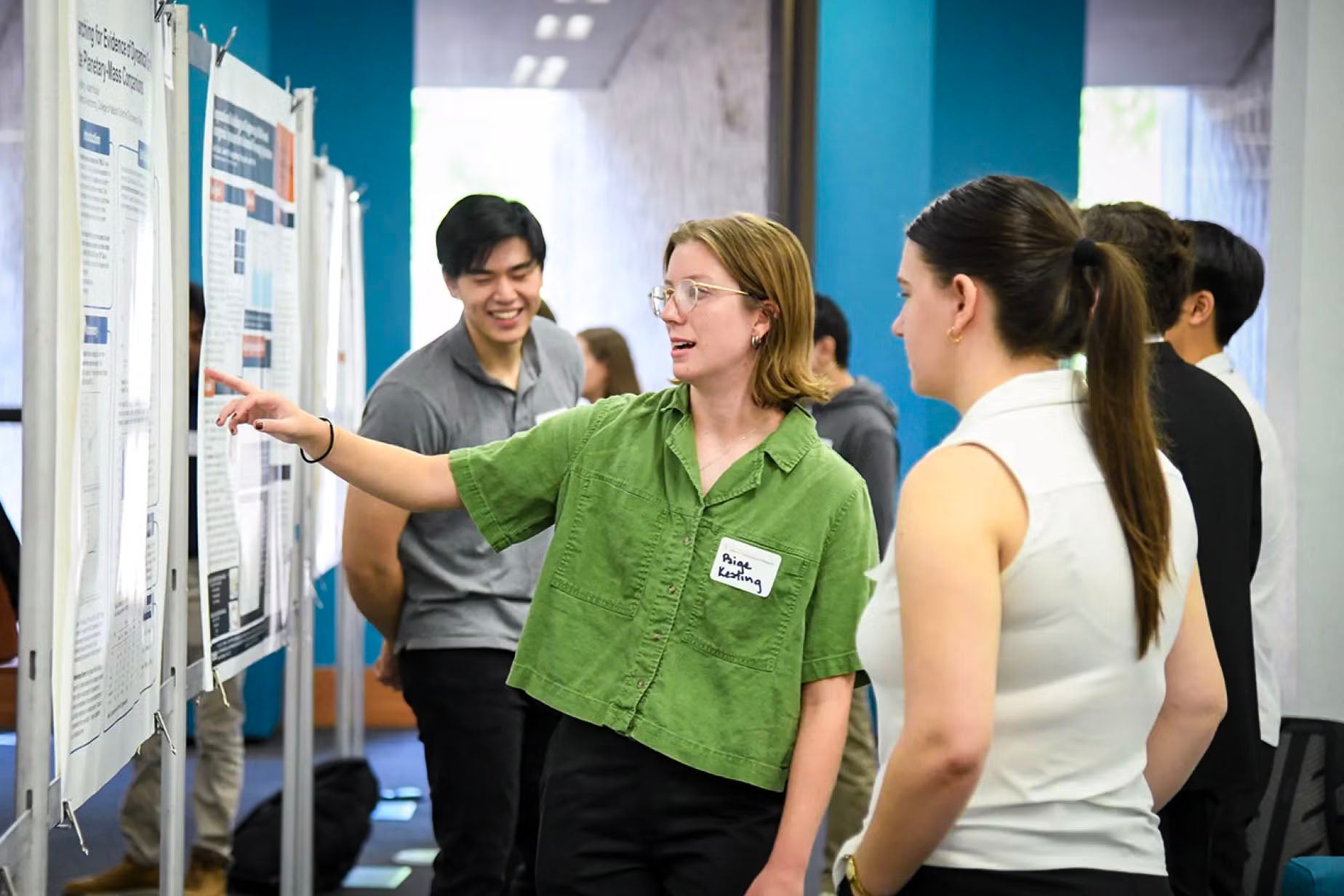 A student wearing a green shirt points to a research poster while discussing her project with another student at an academic poster session. Other students and attendees are visible in the background, engaged in similar presentations.