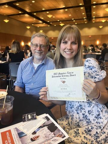 A smiling student, Jane Anderson, holds up a certificate for the 2024 Signature Course Information Literacy Award (First Place) while seated at a table next to Dr. Adam Rabinowitz at a recognition event. Other attendees are visible in the background of the large banquet-style room.
