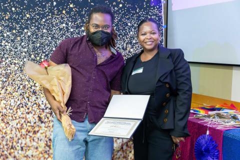 Poet KB Brookins stands next to Associate Professor Simone Browne at an award ceremony. Brookins, wearing a maroon shirt and light blue jeans, holds a bouquet of flowers in one hand and an open certificate in the other. Browne, dressed in a black suit, smiles beside them. The backdrop features a festive starry pattern, and a nearby table is decorated with colorful items, including rainbow flags and party props.