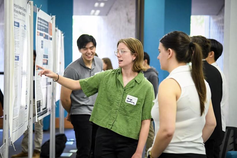 A student wearing a green shirt points to a research poster while discussing her project with another student at an academic poster session. Other students and attendees are visible in the background, engaged in similar presentations.