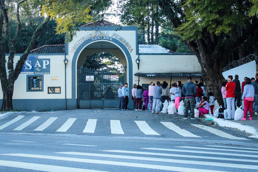 People stand in line outside the entrance gate of the Penitenciária Feminina de Santana, São Paulo’s largest women’s prison, holding bags likely filled with items for incarcerated loved ones. A sign for the São Paulo State Department of Penitentiary Administration (SAP) is visible on the prison wall. Trees frame the scene and a crosswalk leads to the gate.
