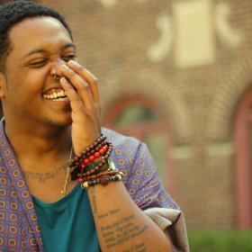 Image of Danez Smith. Man with hand stifling laughter, brick building in the background.