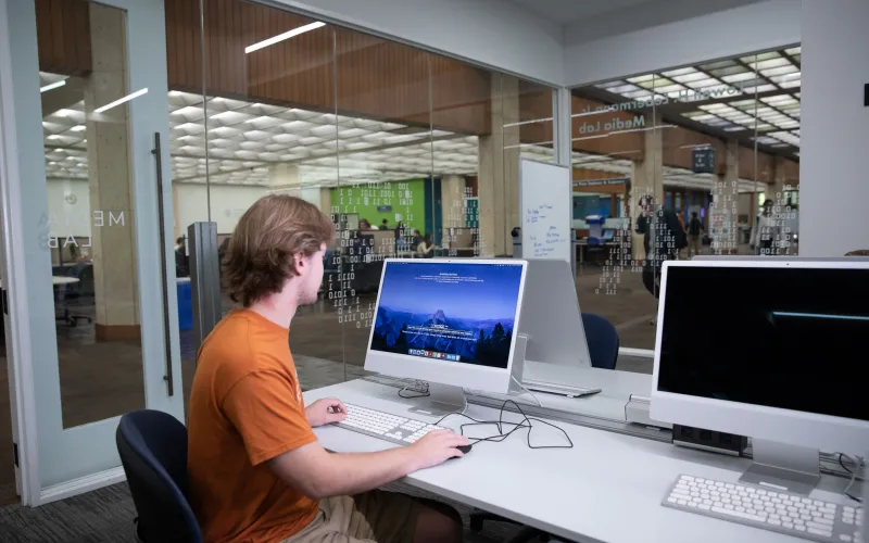 A student in a burnt orange shirt looking at a desktop monitor in a computer lab