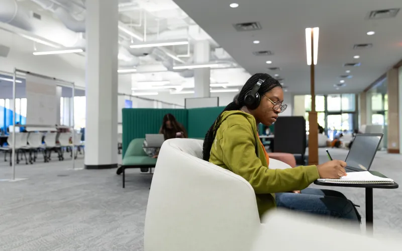 A photo of a student sitting in a comfy chair with headphones on writing something in a notebook