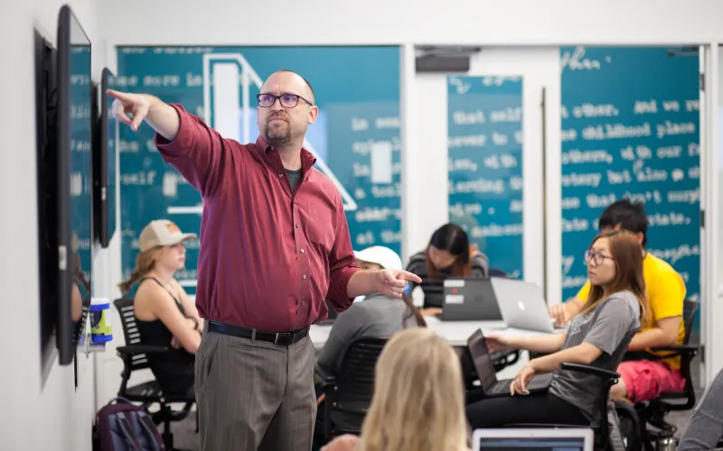 An instructor in a button down shirt pointing at a monitor standing front of a students