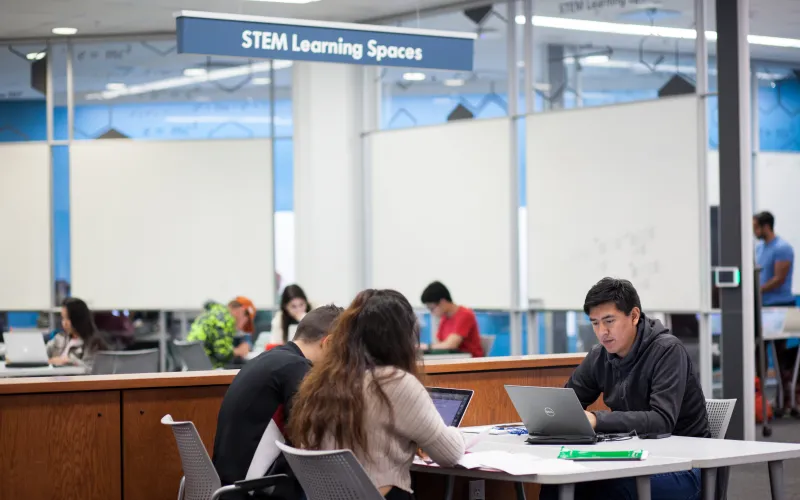Three students sitting at a table together looking at their laptops in the UFCU Room