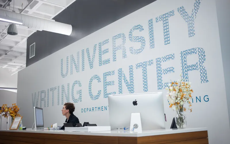 The front desk of the University Writing Center, one person is sitting at the desk looking at a computer.